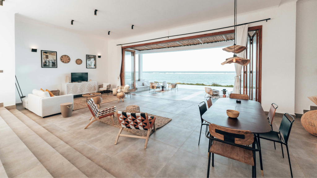 A spacious luxury living and dining area with neutral décor, natural textures, and large open sliding doors overlooking a bright ocean view. Modern furniture, woven accents, and abundant natural light create an elegant coastal atmosphere.