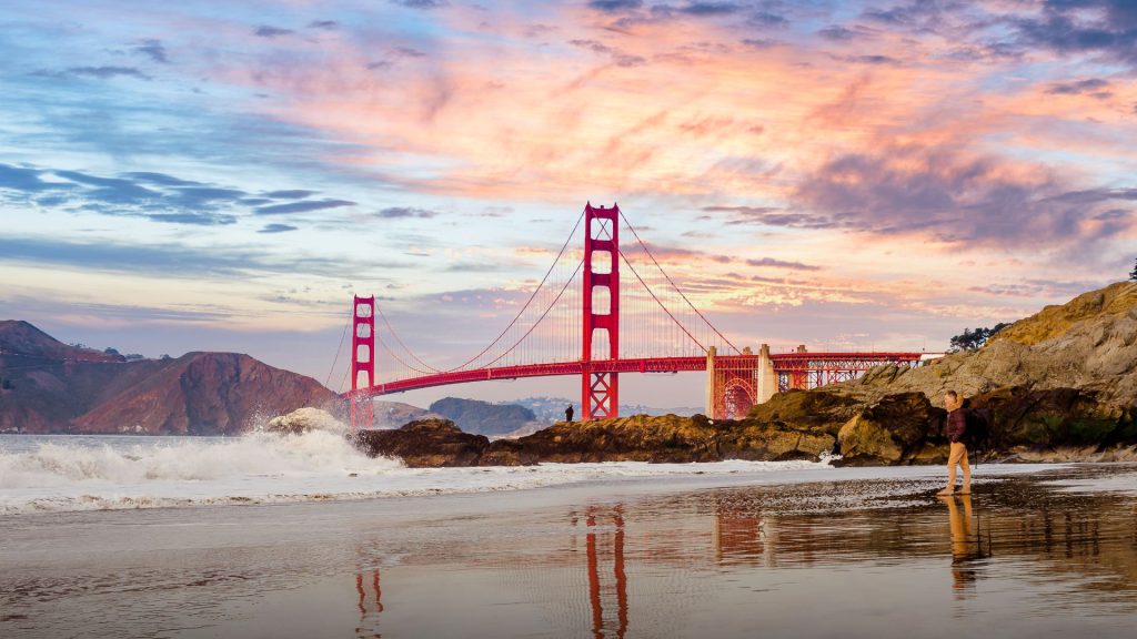 The Golden Gate Bridge at sunset viewed from a sandy beach with a person standing near the crashing waves.