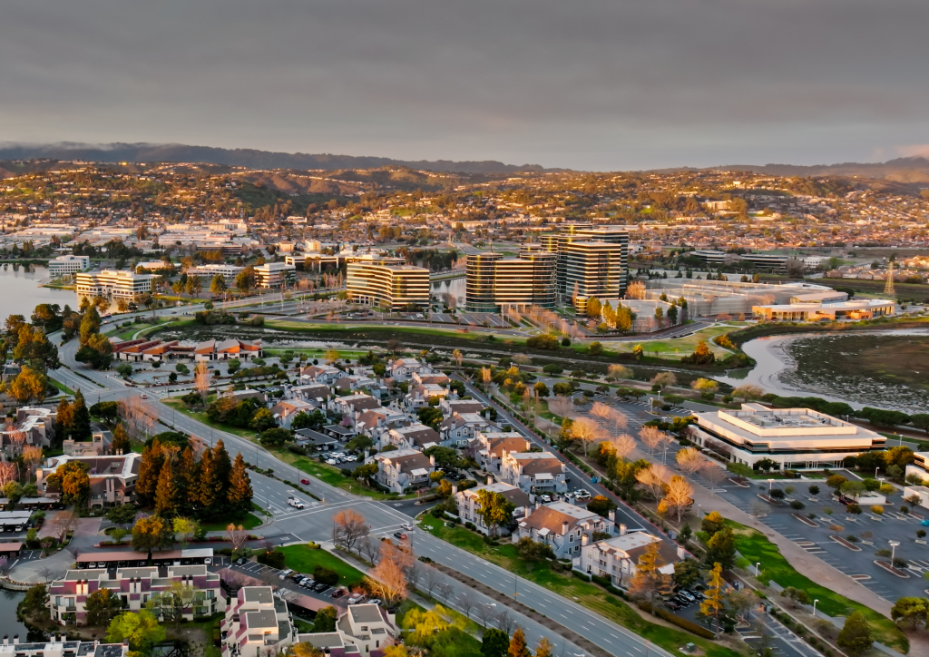 A wide aerial view of Silicon Valley showing dense suburban neighborhoods, tech office parks, and the rolling green hills of the Bay Area.
