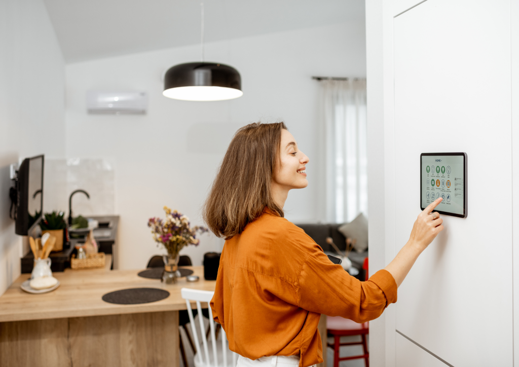 A woman using a wall-mounted smart home tablet to control lighting and security in a bright, modern kitchen.